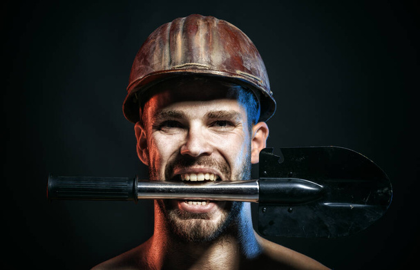 Těžký průmysl a těžba. Mužský dělník, horník v bezpečném tvrdém klobouku s lopatou v zubech. Detailní portrét seriózního profesionálního stavitele v ochranné přilbě s rýčem. Inženýr v klobouku - Fotografie, Obrázek