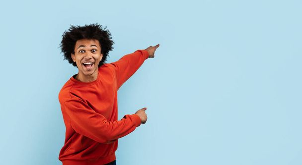 Un hombre negro entusiasta en un suéter rojo apunta con una mirada sorprendida sobre un fondo azul - Foto, imagen