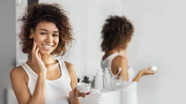 Chica afro feliz aplicando crema en su cara, de pie en el baño, reflejándose en el espejo. - Foto, Imagen