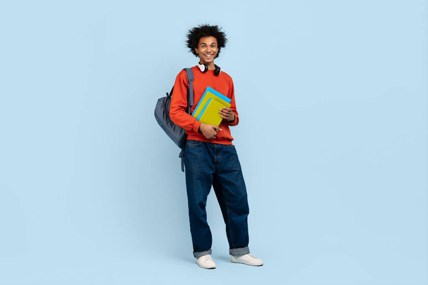 Casual dressed young african american man holding books with a backpack on a blue background - Foto, immagini
