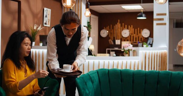 Aziatische ober brengt koffie naar de gasten in de lounge, wachtend om te worden gebeld aan de receptie voor kamer check-in. Vrouw die aankomt in het resort en ontspannen met een drankje voor de registratie, vriendelijk personeel. - Foto, afbeelding