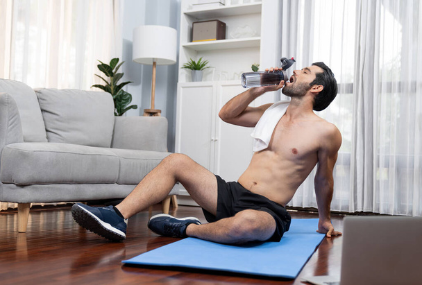 Athletic and sporty man drinking water on fitness mat after finishing home body workout exercise session for fit physique and healthy sport lifestyle at home. Gaiety home exercise workout training. - Fotó, kép