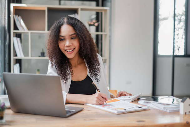 Une jeune femme d'affaires souriante travaille sur des documents financiers avec une calculatrice et un ordinateur portable à un bureau ensoleillé. - Photo, image