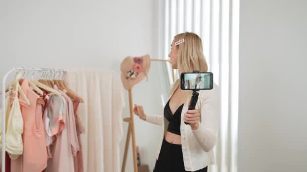 Joven creador de contenido social medial mujer hacer video de moda utilizando palo selfie. Blogger sonríe a la cámara mientras hace que la ropa en línea persuasiva vlog de la venta a la audiencia o al seguidor. Blithe. - Imágenes, Vídeo