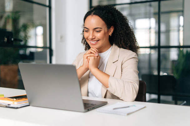 Happy brazilian or hispanic curly haired business woman, company ceo, recruitment manager, sitting at a work desk in a modern creative office, working in a laptop, studying information, smiling - Photo, Image