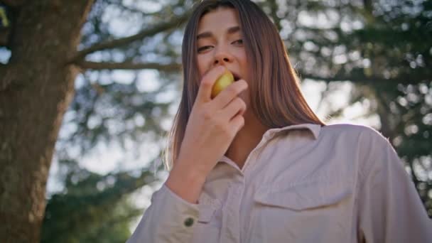 Woman eating apple park standing at sunlight closeup. Smiling beautiful lady delighting in taste of fresh fruit on sunny nature enjoy healthy snack outdoors. Happy girl chewing in front green foliage. - Footage, Video