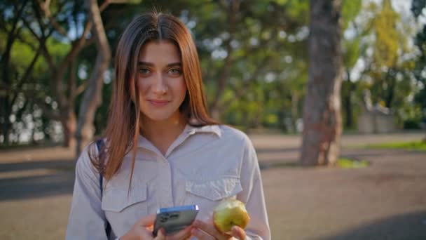 Αρραβωνιασμένη γυναίκα ψάχνει smartphone κρατώντας μήλο στο πράσινο ηλιόλουστο πάρκο closeup. Πορτρέτο της ευτυχούς κυρία κλήση αριθμό τηλεφώνου στην όμορφη φύση. Θερινός ελεύθερος χρόνος με τεχνολογία που αντικατοπτρίζει τον περιστασιακό τρόπο ζωής. - Πλάνα, βίντεο