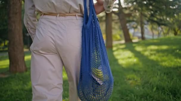Vědomá žena nesoucí ekologicky použitelnou nákupní tašku v zeleném slunečném parku. Nenáročný nakupující, který drží ekologické produkty, přijímá udržitelnost ve venkovním prostředí. Detailní dívka ruka uchopení módní eko-šetrné příslušenství - Záběry, video