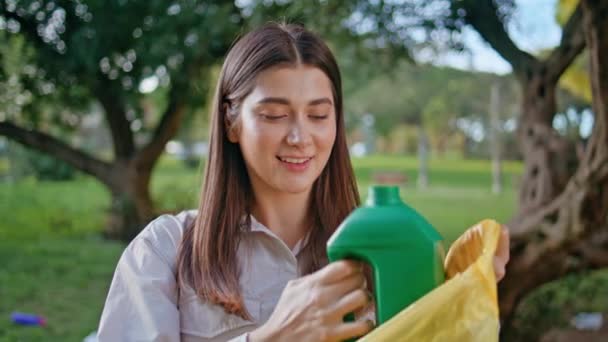 Mulher sorridente jogando garrafa de plástico no saco de reciclagem irradiando felicidade eco-friendly no parque. Fechar voluntariado alegre mostrando gestão sustentável de resíduos. Retrato de ambientalista feminina. - Filmagem, Vídeo