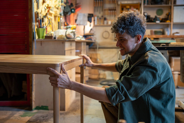 Εργαστήριο ξυλουργικής. Νεαρός επαγγελματίας ξυλουργός ελέγχει την ποιότητα της εργασίας, εξετάζει το ξύλινο τραπέζι μετά το βερνίκωμα, χαίρεται για την τέλεια εκτέλεση της εργασίας. Κατασκευή εξατομικευμένων επίπλων. - Φωτογραφία, εικόνα