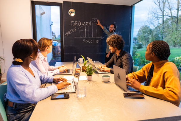 Em um escritório moderno e bem iluminado, um grupo de profissionais focados se senta em torno de uma mesa trabalhando em laptops, enquanto um membro da equipe apresenta entusiasticamente estratégias de crescimento em um quadro-negro, incorporando startup - Foto, Imagem