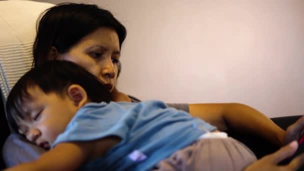 Mãe olhando para smartphone sentado no sofá à tarde com seu bebê em seu peito e lendo algo. - Filmagem, Vídeo