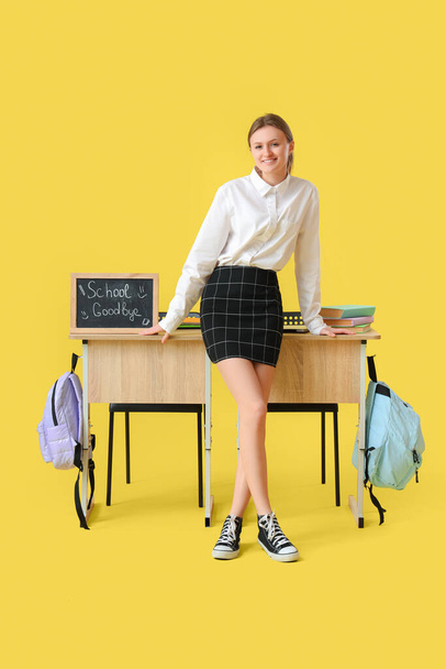 Estudiante cerca de escritorios con pizarra con texto GOODBYE SCHOOL sobre fondo amarillo. Concepto de fin de escuela - Foto, imagen