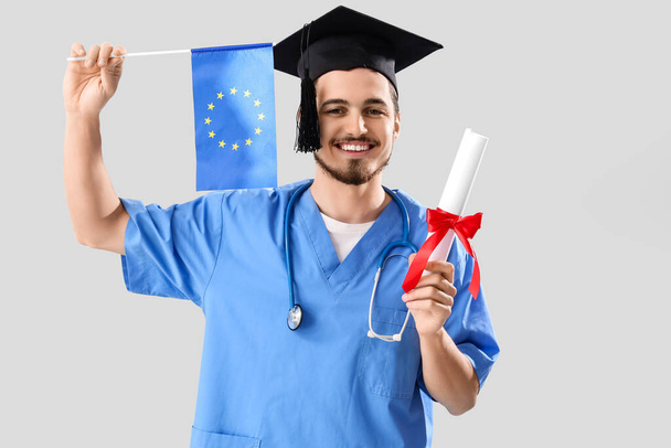 Άρρεν φοιτητής ιατρικής με δίπλωμα και σημαία ΕΕ σε λευκό φόντο - Φωτογραφία, εικόνα