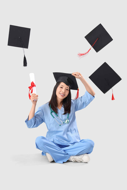 Asian female medical graduate student with diploma and falling mortar boards on white background - Photo, Image
