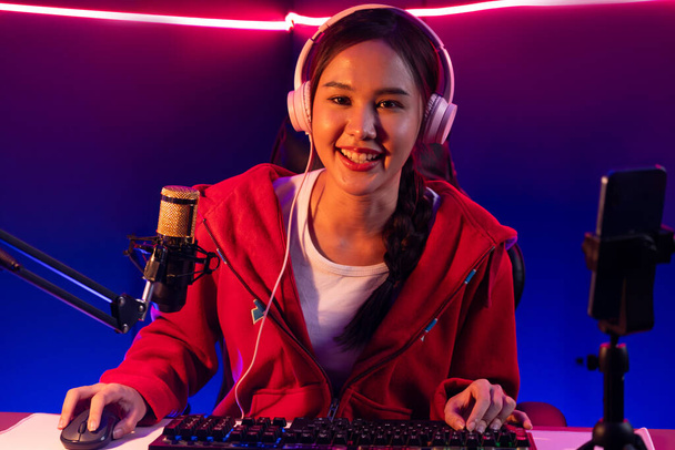 Host channel of smiling beautiful Asian girl streamer playing online game wearing headphone talking with viewers media online recording phone. Esport skilled team players in neon blue room. Stratagem. - Фото, зображення