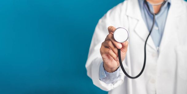 Hand van een arts die een stethoscoop vasthoudt terwijl hij op een blauwe achtergrond staat. Een close-up foto. Ruimte voor tekst. Medisch, behandelings- en gezondheidsconcept. - Foto, afbeelding