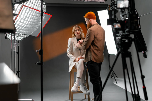 Ein Mann unterhält sich mit einer Frau auf einem Hocker vor einer Kamera für ein Unterhaltungsfernsehprogramm. Zur Bühnenausstattung gehören ein Stativ und ein stylischer Modeanzug - Foto, Bild