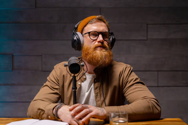 Een man met een baard, een bril en een koptelefoon, zit aan een tafel met een microfoon voor zich, klaar om op te nemen. Podcast studio concept - Foto, afbeelding