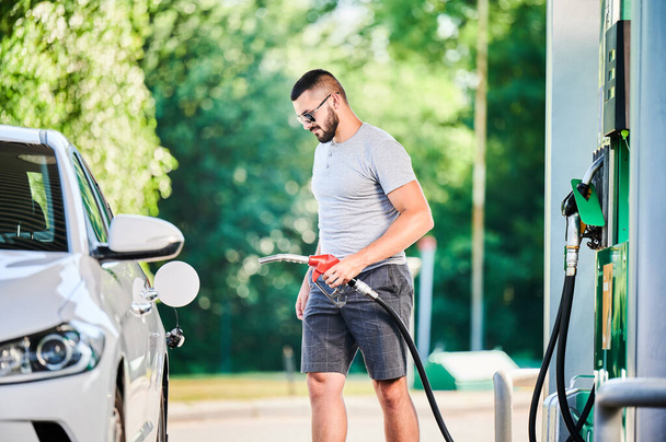 Benzin pompalı bir sürücü benzin deposunu dolduruyor. Lüks beyaz otomobiline yakıt dolduran kendinden emin bir adam. Modern benzin istasyonunda günlük kıyafetler ve güneş gözlüğü takan bir adam.. - Fotoğraf, Görsel