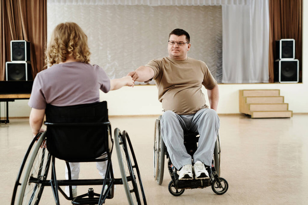 Зрелый мужчина и неузнаваемая женщина в инвалидных колясках практикуют танец в современной просторной студии - Фото, изображение