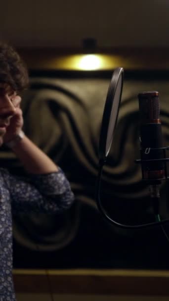 Spokojny wokalista w słuchawkach nagrywający utwór muzyczny w profesjonalnym studio nagraniowym. Mężczyzna śpiewający piosenkę do mikrofonu ze słuchawkami na głowie na tle dźwiękochłonnej gumy piankowej - Materiał filmowy, wideo