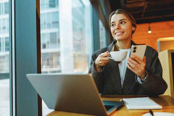 Vrouwelijke manager leest bericht op telefoon en werkt op laptop in café. Begrip "werken op afstand" - Foto, afbeelding