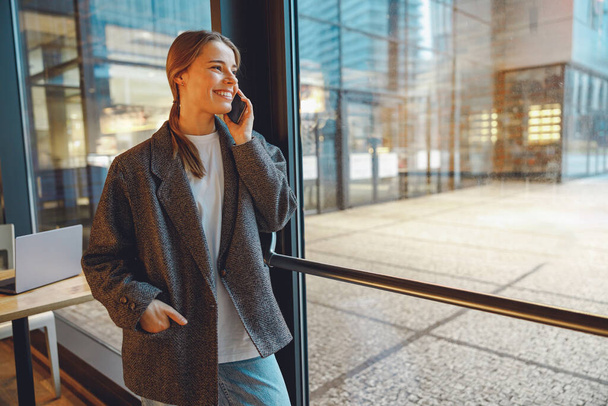 Usmívající se podnikatelka mluví telefon s kolegy, zatímco stojí v kavárně u okna - Fotografie, Obrázek