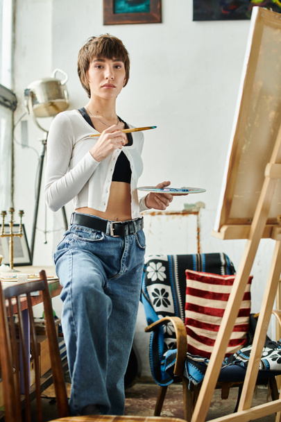 A woman holding a paintbrush, standing in front of an easel. - Photo, Image