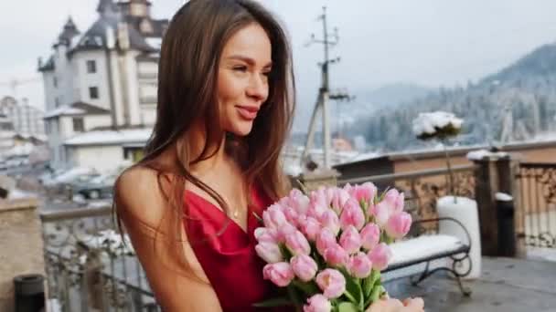 Retrato de una mujer bonita con un vestido rojo con tulipanes frescos en las montañas. La mujer posa para la cámara con un ramo de flores - Imágenes, Vídeo