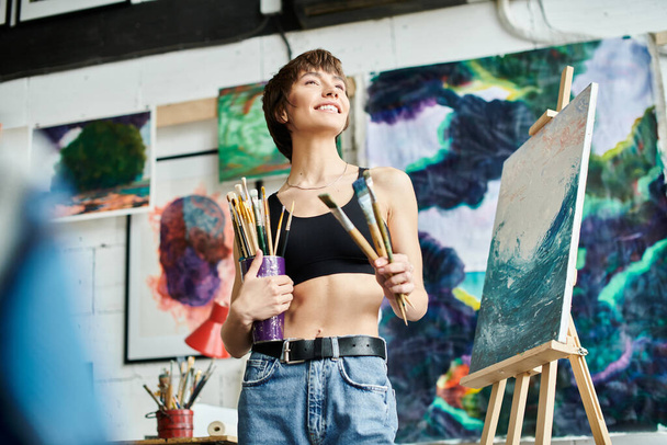 A woman with a paintbrush stands before a painting. - Photo, Image