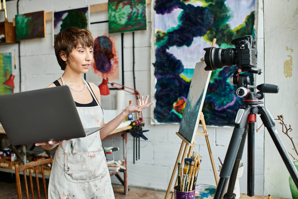 Γυναίκα στο στούντιο τέχνης κρατώντας ένα φορητό υπολογιστή, που περιβάλλεται από δημιουργικότητα. - Φωτογραφία, εικόνα