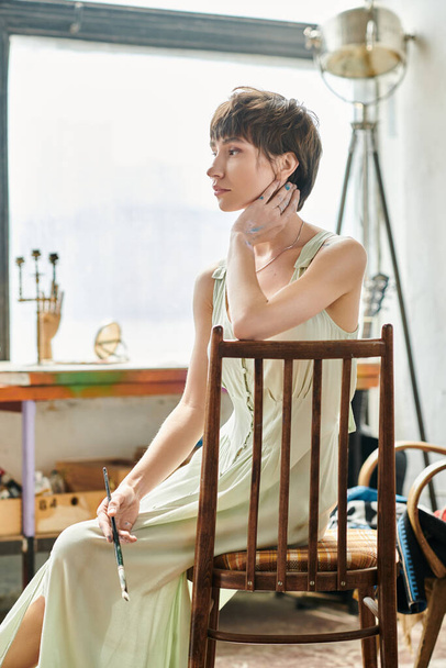 Μια γυναίκα με μακρύ φόρεμα κάθεται με χάρη σε μια καρέκλα.. - Φωτογραφία, εικόνα