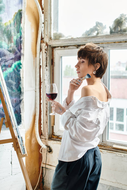 Una mujer está de pie junto a una ventana, sosteniendo una copa de vino. - Foto, Imagen