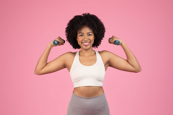 Una mujer afroamericana feliz con cabello natural sostiene pesas, mostrando fitness sobre un fondo rosa - Foto, Imagen