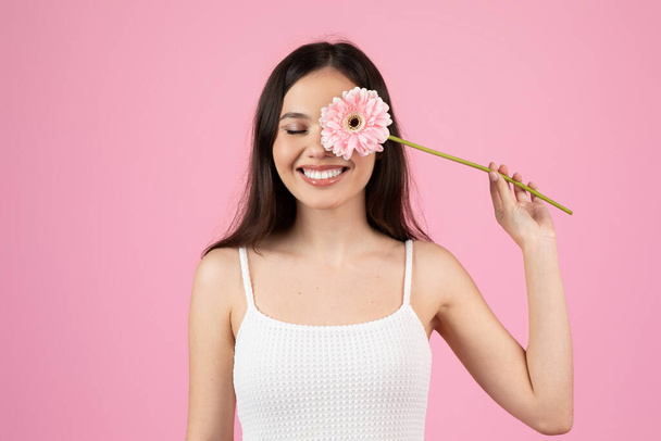 Una joven jovial pone juguetonamente una flor rosa gerbera cerca de su cara sobre un fondo rosa de estudio - Foto, imagen