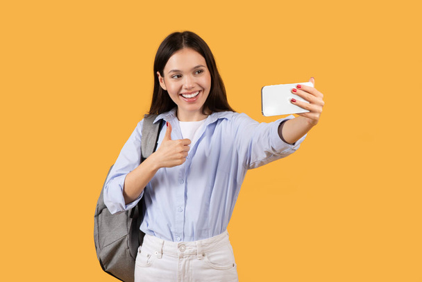 A cheerful young woman with a backpack takes a selfie, smiling joyfully and showing a thumbs up gesture on a yellow background - Photo, Image