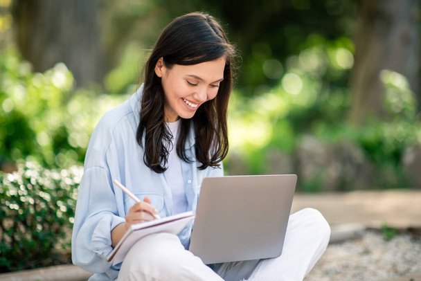 Een toegewijde jonge studente gebruikt haar laptop om buiten in het park te studeren, aantekeningen te maken in een notitieblok - Foto, afbeelding