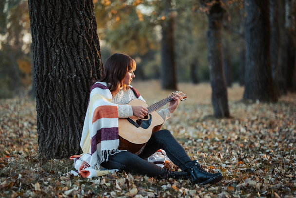 森の中の涼しい秋の夕方,暖かい服を着た若い女の子が葉の上に座って一人でギターを弾きました. 楽器と自分の思考で時間を過ごすという概念 - 写真・画像