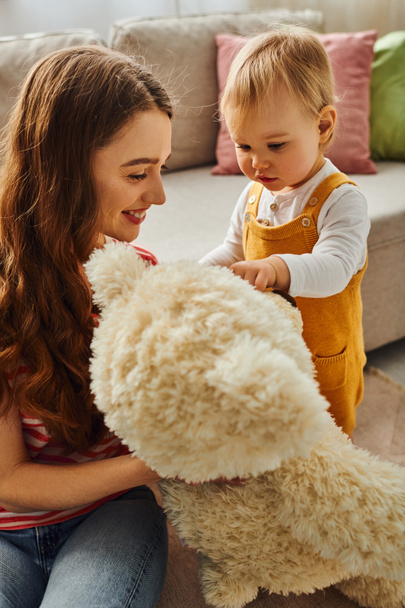Una giovane madre tiene amorevolmente un animale di peluche accanto alla sua bambina a casa, creando un momento commovente di affetto.. - Foto, immagini