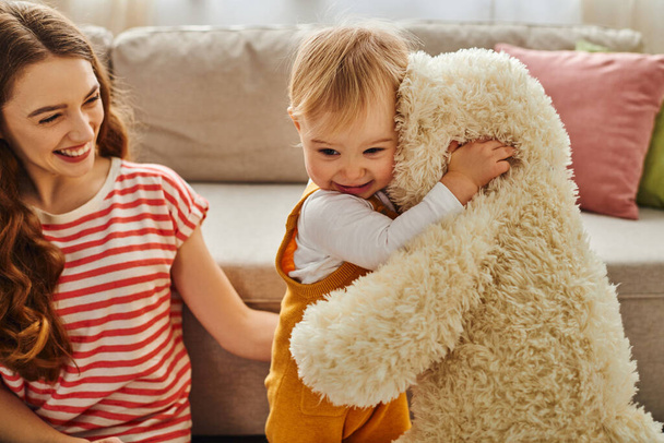 Une jeune mère tient tendrement son bébé tout en embrassant un ours en peluche dans une démonstration chaleureuse d'amour à la maison. - Photo, image