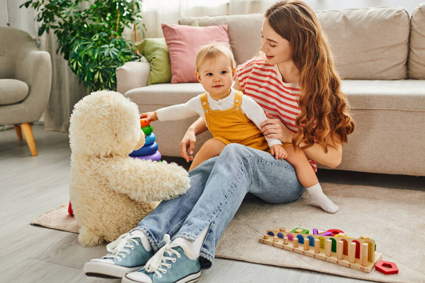 Een jonge moeder zit op de vloer naast haar peuter dochter en een teddybeer, genietend van een rustig moment samen. - Foto, afbeelding