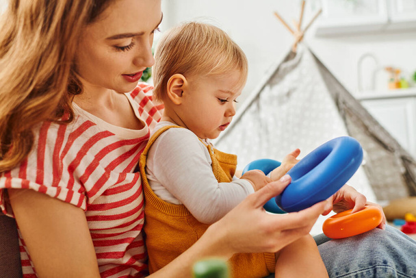 Uma jovem mãe segura sua filha bebê enquanto brinca com um brinquedo colorido, criando um momento de amor e riso. - Foto, Imagem