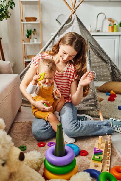 Una joven madre está jugando felizmente con su hija pequeña en una habitación acogedora, construyendo una conexión amorosa y alegre. - Foto, imagen