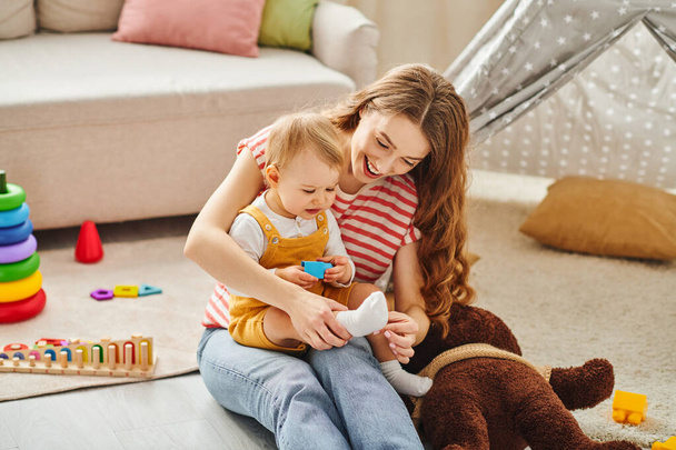 Молода мати сидить на підлозі, граючи зі своєю дочкою малюка в сердечний момент зв'язку і радості. - Фото, зображення