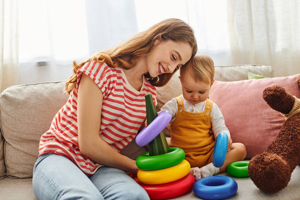 Una madre joven y su hija pequeña riendo y jugando en un sofá acogedor, creando valiosos recuerdos juntos. - Foto, imagen
