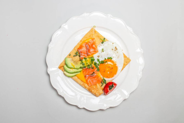Detailní záběr krepu s uzeným lososem, avokádem, vejcem a rajčaty na bílém talíři. čtvercový tvar, elegantní prezentace. - Fotografie, Obrázek