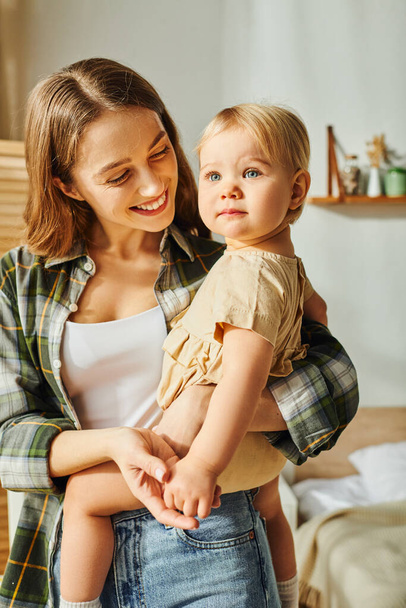 Mladá matka kolébá svou batolata dceru v náručí, sdílí moment lásky a spojení doma. - Fotografie, Obrázek