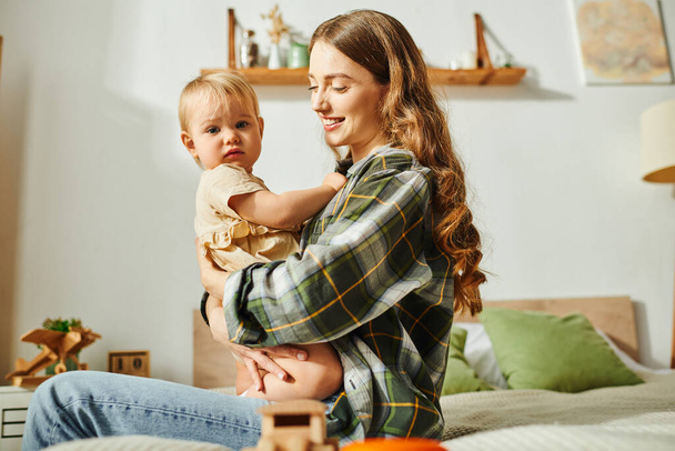 Mladá matka sedící na posteli, něžně držící svou dcerku v náručí a ctící spolu vzácný okamžik. - Fotografie, Obrázek