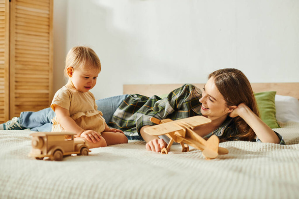 Eine junge Mutter liegt auf einem Bett neben ihrer kleinen Tochter und teilt einen heiteren und herzerwärmenden Moment der Verbundenheit. - Foto, Bild